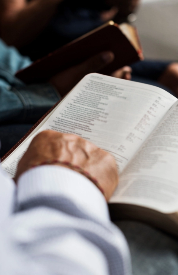 A person wearing a striped shirt reads an open Bible close to the camera, with another person holding a Bible in the background.