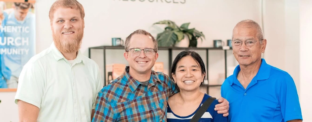 Four people stand together smiling in a room with a shelf and plants in the background. The wall behind them displays the words "AUTHENTIC CHURCH COMMUNITY" partially visible, reflecting their involvement with Church Ministries in Raleigh NC.