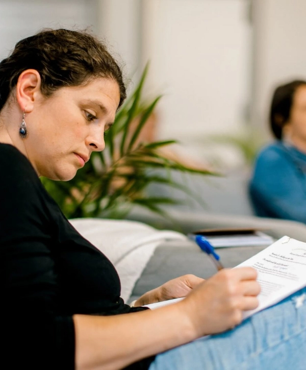 A woman in a black top and blue jeans is sitting and writing on a piece of paper with a blue pen. She appears focused and is surrounded by the soft, blurred indoor background of the Local City Church in Raleigh, NC.