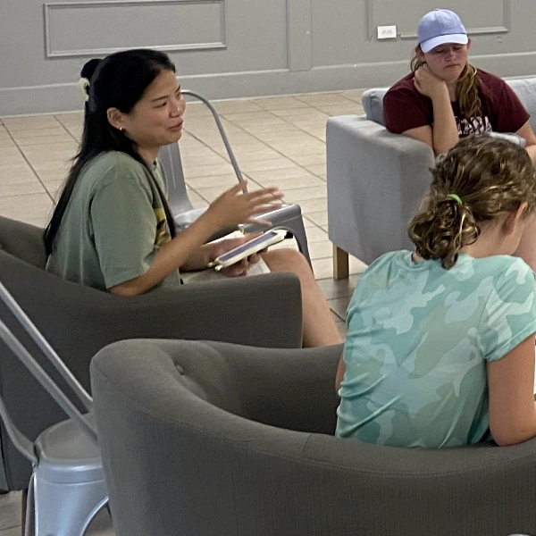Three individuals sit on chairs having a discussion in a gray-walled room. One person is speaking, the second person is writing, and the third person is listening with a hand on their chin.