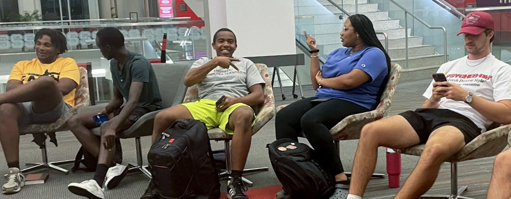 Five people sitting in chairs arranged in a semi-circle, engaged in conversation and activities. Three people are talking and smiling, while one person looks at their phone and another observes—reminiscent of the welcoming atmosphere often found at Church Ministries in Raleigh NC.
