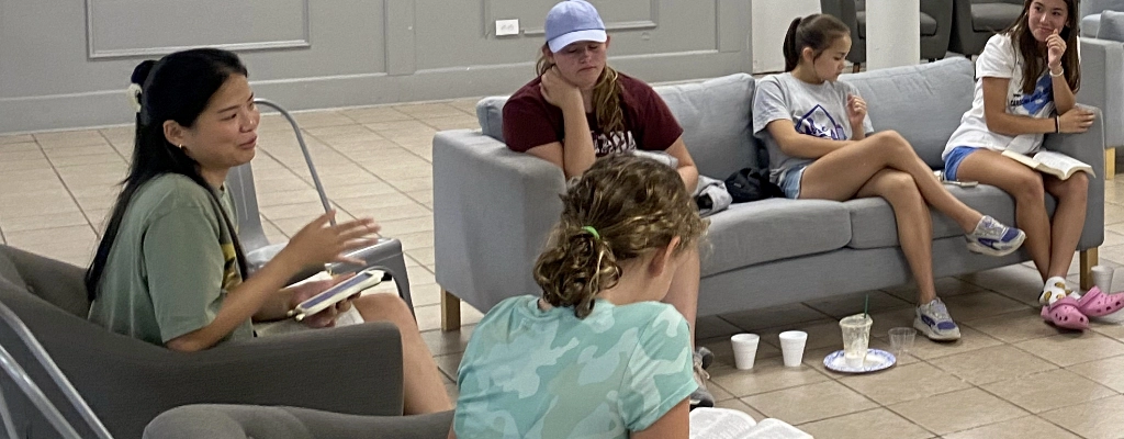 A group of five young individuals from Church Ministries in Raleigh NC sit on couches and chairs in a casual setting, reading and engaging in conversation.