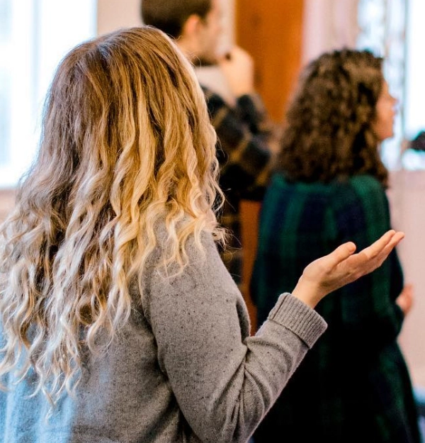 A person with long wavy hair raises their hand, facing away, in the Local City Church in Raleigh, NC. Other people, slightly out of focus, are seen in the background.