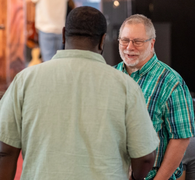 Two men are engaged in conversation indoors at the Christian Church in Raleigh. One man is facing the camera, smiling, while the other has his back to the camera.