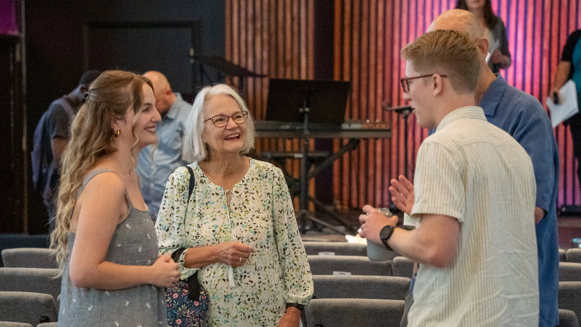 Four people are engaged in conversation in a room with rows of chairs and a music setup in the background. Additional people are partially visible further back.